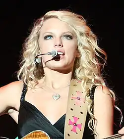 A blonde woman wearing a dark shirt is playing the guitar and singing